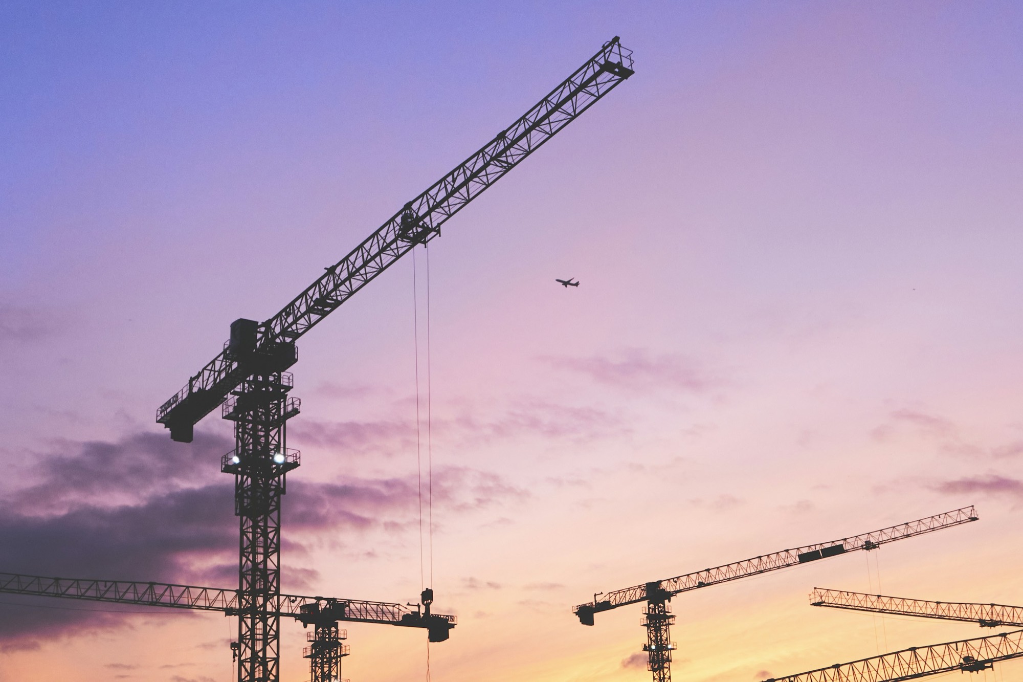Gru da cantiere stagliate contro un cielo al tramonto, con un aereo in volo sullo sfondo